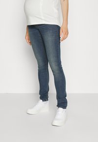 Mørkeblå gravidejeans med slim fit, glatt denimtekstur og subtil fading, paret med hvite joggesko. Hvit topp delvis synlig.