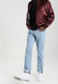 Blouson aviateur en satin bordeaux, pull noir, jean bleu clair et baskets blanches avec revers et détails subtils de texture.
