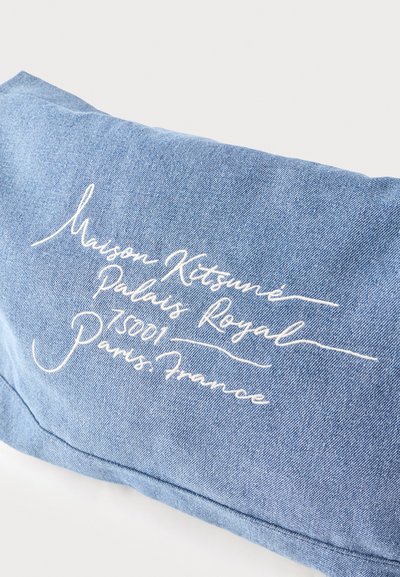 Coussin en denim bleu avec un texte en cursive blanche brodé sur le devant indiquant "Maison Kitsuné Palais Royal 75001 Paris France".