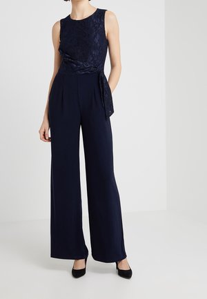 Woman wearing sleeveless navy lace top with side tie and matching wide-leg trousers, black pointed heels, against plain background.