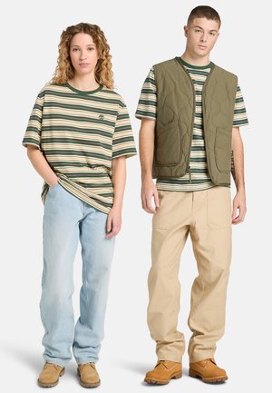 Młoda kobieta w zielono-beżowej koszulce w paski i jasnych dżinsach, młody mężczyzna w pasującej koszulce w paski, zielonej pikowanej kamizelce, beżowych spodniach i brązowych butach.