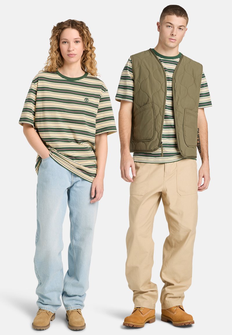 Jeune femme en tee-shirt rayé vert et beige avec un jean clair, jeune homme en tee-shirt rayé assorti, gilet matelassé vert, pantalon beige et bottes marron.