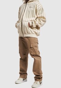 Person som bär en beige zip-up hoodie med vågig linjemönster, bruna cargobyxor och krämfärgade sneakers med svart swoosh-logotyp.