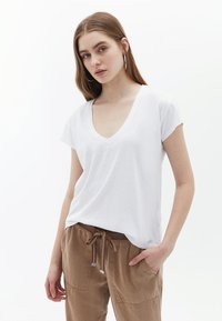 T-shirt bianca con scollo a V realizzata in tessuto leggero, con maniche corte e vestibilità ampia, abbinata a pantaloni marroni con cordoncino e tasche.