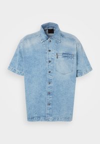 Camisa de mezclilla de manga corta en azul claro, con un cierre de botones en la parte delantera, cuello y un bolsillo en el pecho con cierre de cremallera. Textura ligeramente desgastada.