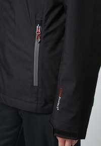 Gros plan sur une manche de veste noire avec l’étiquette "Clima Protect", un poignet en velcro et une poche zippée avec des tirettes rouges et grises.