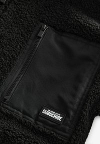Détail d'une veste en polaire noire avec une couche extérieure texturée et une poche zippée noire lisse portant une étiquette indiquant "SUB5CRIBE."