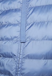 Jasnoniebieska kamizelka puchowa z pikowanymi sekcjami, centralnym zamkiem błyskawicznym i pętelką z materiału do zapięcia. Gładki, błyszczący materiał o minimalnej teksturze.