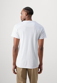 Camiseta blanca de algodón con mangas cortas, escote redondo y un dobladillo trasero ligeramente más largo, combinada con pantalones beige. Diseño liso, sin patrones.
