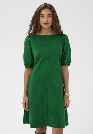Žena s kučeravými hnedými vlasmi, ktorá má na sebe zelené šaty po kolená s nadýchanými krátkymi rukávmi, stojí pred jednofarebným svetlým pozadím.