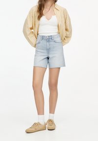High-waisted light blue denim shorts with frayed hem, paired with a white tank top and a beige striped button-up shirt. Beige sneakers and white socks.