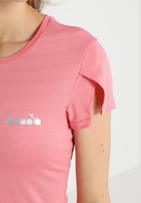 Camisa deportiva rosa con un escote redondo, mangas cortas con un corte único y un logo plateado en el pecho. Diseño de tela texturizada.