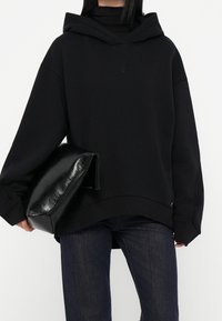 Sudadera negra de gran tamaño con bolsillo tipo canguro y un discreto bordado de logo. Combinada con vaqueros oscuros y un bolso clutch negro con textura.