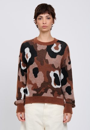 Vrouw met kort donker haar, die een bruin, zwart, wit en roze abstract bloemmotief gebreide trui en lichte broek draagt tegen een effen achtergrond.