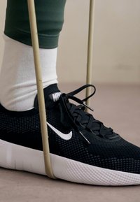 Crna Nike tenisica s teksturiranim gornjim dijelom, bijelim logotipom i bijelim potplatom, u paru s crno-bijelim čarapama i zelenim sportskim tajicama.