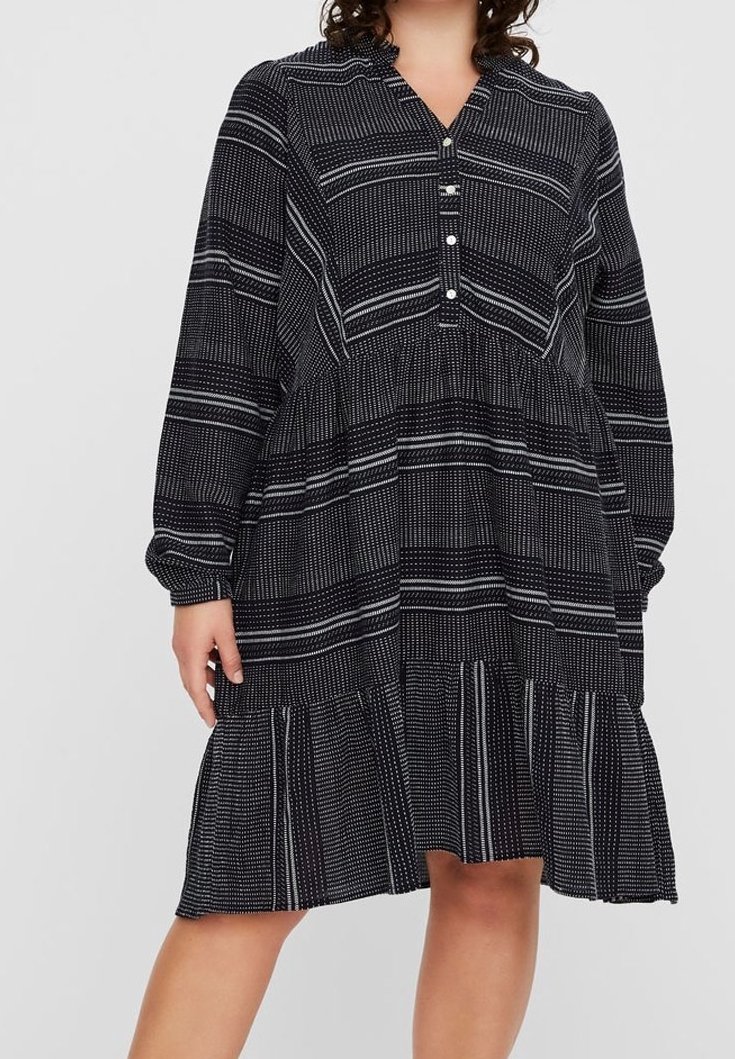 Femme portant une robe noire ample avec des motifs à pois blancs et rayures, manches longues, boutons sur la poitrine et un ourlet de jupe à volants.