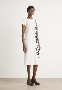 Abito bianco al ginocchio con design aderente, maniche corte a cappuccio e motivo floreale nero su un lato. Indossato con sandali neri con tacco.