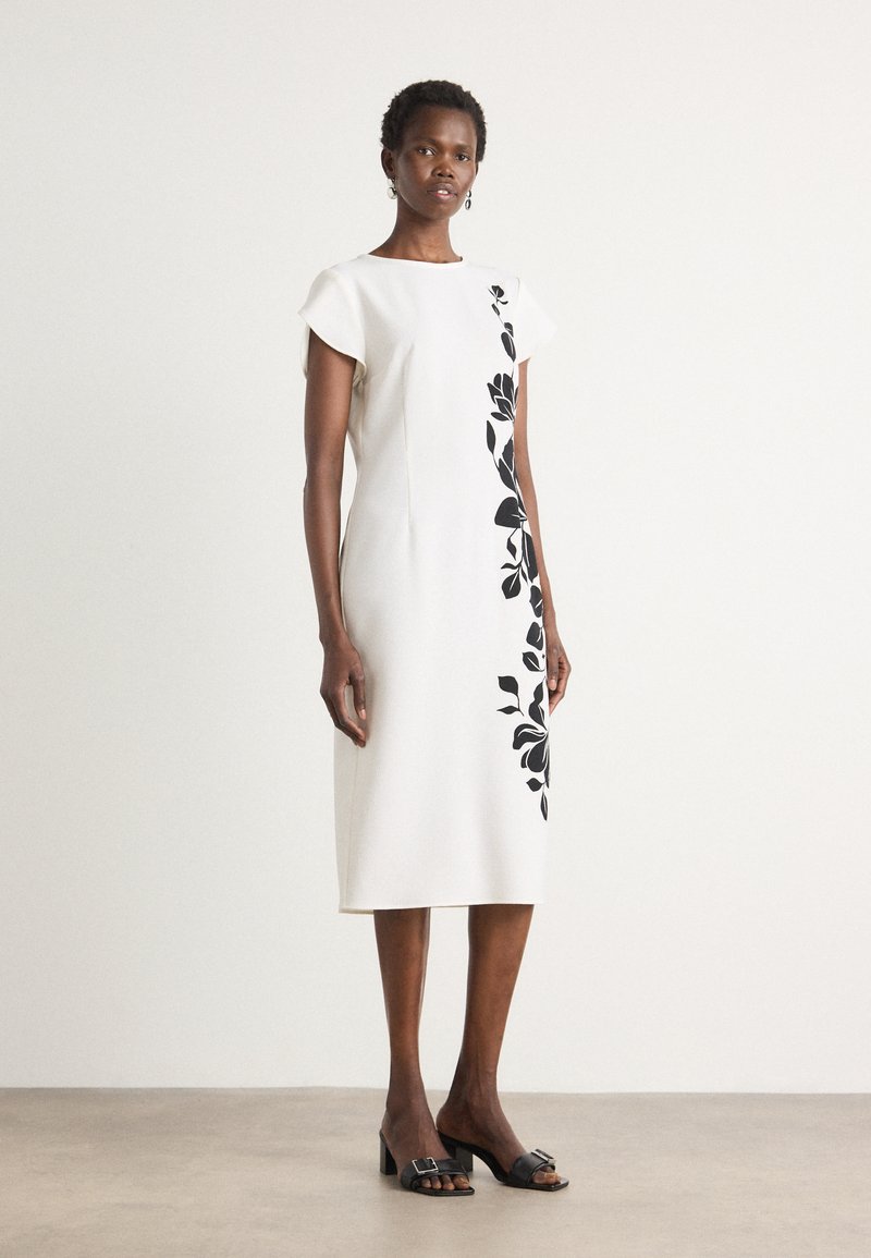White knee-length dress with a fitted design, short cap sleeves, and black floral pattern along one side. Worn with black heeled sandals.