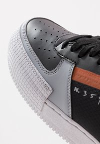Svart och grå sneaker i perforerat läder, med texturerade gummidetaljer, vit sula och ett orange sträck med numerisk detalj.