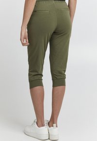 Person med olivengrønne, cropped bukser med elastiske manchetter og hvite sneakers, vist fra baksiden mot en enkel bakgrunn.