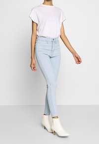 Žena v světlomodrých vysokých skinny džínách, bílém tričku s krátkým rukávem a bílých kotníkových botách, stojící na jednotném pozadí.