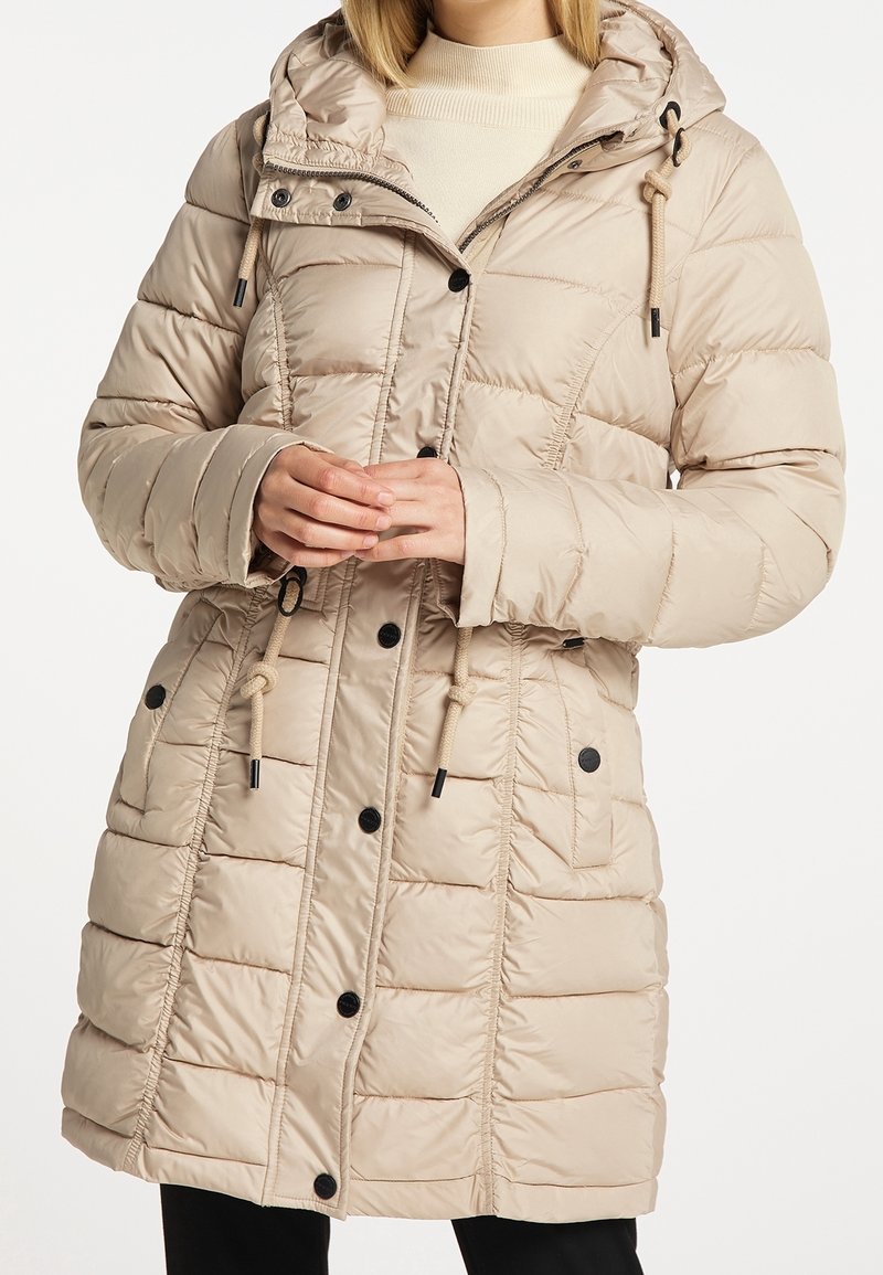 Doudoune matelassée beige avec capuche, boutons noirs, cordons de serrage et poches avant, portée sur un pull crème.