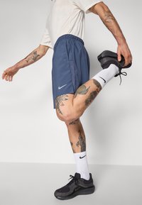 Navy athletic shorts with an elastic waistband, paired with black sneakers and white Nike socks. Tattoos visible on the leg, textured skin.