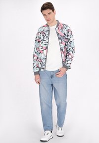 Jachetă bomber colorată cu un model floral, manșete și guler gri cu caneluri, asociată cu un tricou alb, blugi deschiși la culoare și adidași albi.
