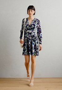 Robe à imprimé floral avec des manches longues, un décolleté en V profond et une taille empire. Les couleurs incluent le violet, le vert et le blanc. Associée à des sandales à lanières.
