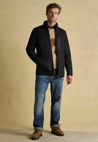 Czarna pikowana kurtka z wysokim kołnierzem, na zamek błyskawiczny, z dwoma przednimi kieszeniami, w połączeniu z beżowym swetrem, niebieskimi dżinsami i brązowymi butami.