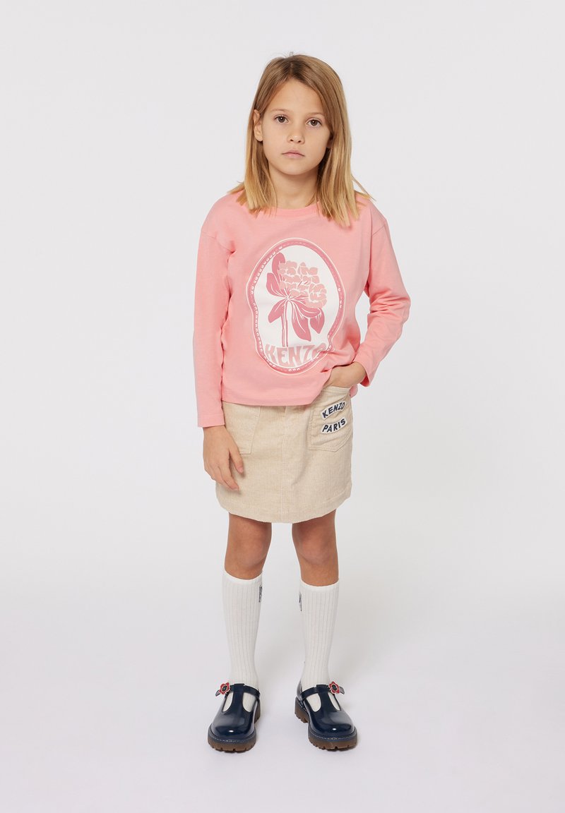 Chemise rose à manches longues avec un graphique floral blanc, jupe en velours côtelé beige, chaussettes blanches montantes et chaussures bleu foncé avec des accents floraux.