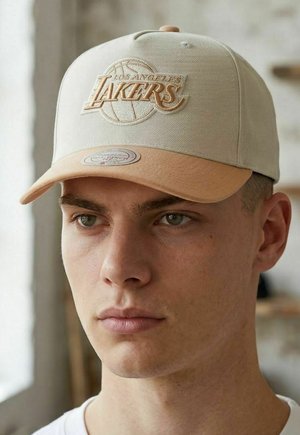 Joven con gorra de Los Angeles Lakers en beige y marrón claro con logo bordado, mirando ligeramente hacia la derecha con una luz interior suave.