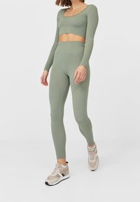 Ribbed, long-sleeve green crop top with a scoop neckline and matching high-waisted leggings, paired with beige athletic shoes.
