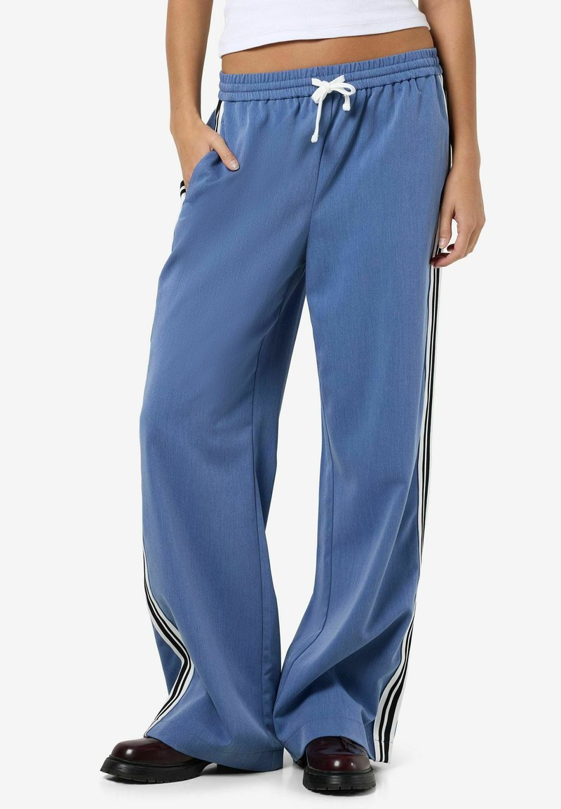 Pantalons larges bleus avec taille à cordon, présentant des accents latéraux rayés noir et blanc et un tissu lisse et texturé.