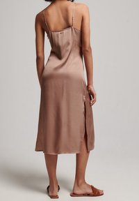 Satin midi kjole i taupe med tynde spaghetti stropper, en sidelange slids og en afslappet pasform. Har en lille knapdetalje bagpå.