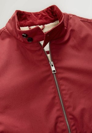 Burgundská bunda s vysokým límcem, která má přední zip, dvě černé knoflíkové zapínání a měkkou krémovou podšívku.
