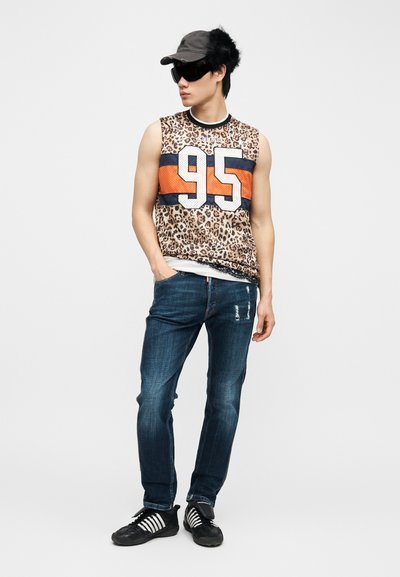 Jeune homme portant un débardeur à imprimé léopard avec le numéro 95, un jean foncé, une casquette noire, de grandes lunettes de soleil noires et des baskets noires à rayures blanches.