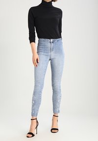 Höga ljusblå jeans med blommönster broderat på underbenen, i kombination med en svart polotröja och svarta klackskor med remmar.