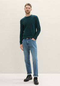 Suéter de un verde oscuro con ribetes, combinado con jeans de un azul claro y zapatos negros con cordones. El conjunto presenta un diseño ajustado y texturas sutiles.