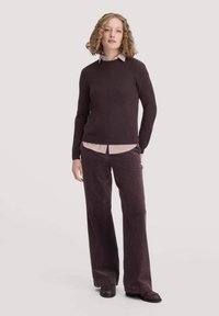 Bordeauxroter Pullover mit langen Ärmeln über einem hellrosa und weißen gestreiften Hemd, kombiniert mit weiten Cordhosen in passendem Bordeauxrot.