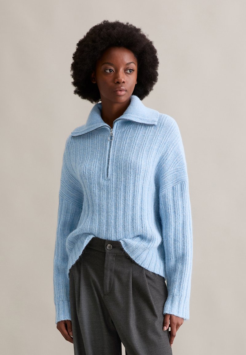 Hellblau geripptes Strickpullover mit einem Reißverschlusskragen, seitlichen Schlitzen und lässigen Ärmeln, kombiniert mit grauen Hosen.