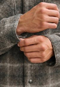 Graues, strukturiertes Wollhemd mit einem Knopfdetail an den Manschetten. Der Stoff hat ein leicht unscharfes Aussehen mit einem dezenten Muster. Hände, die den Ärmel anpassen.