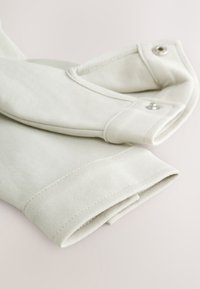 Manches de vêtement couleur crème pliées avec poignets côtelés et boutons-pression, posées à plat sur une surface claire.