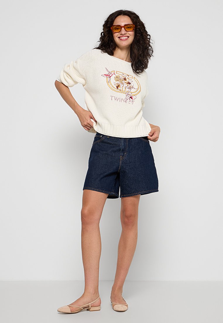 Witte, gestructureerde trui met bloemenborduurwerk; gecombineerd met donkere spijkerbroekshorts; model draagt getinte zonnebril en beige flats.