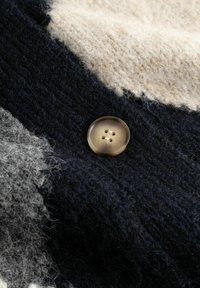 Nahaufnahme eines beigen Knopfs auf einem dick gestrickten Stoff mit schwarzen, weißen und grauen Farbfeldern und einer flauschigen Textur.