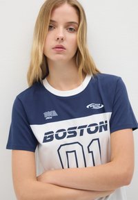 Camiseta azul marino y blanca con "BOSTON 01" impreso en negrita. Presenta un cuello redondo, mangas cortas y detalles gráficos en el pecho.