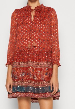 Femme portant une robe rouge fleurie, manches longues, col à nouer avec pompons et ourlet à motifs au-dessus des genoux.