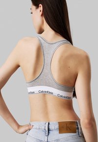 Bralette gris de tirantes en la parte posterior, hecho de una tela suave y texturizada, con una banda blanca que presenta el logo de "Calvin Klein". Combinado con unos pantalones cortos de mezclilla clara de talle alto.