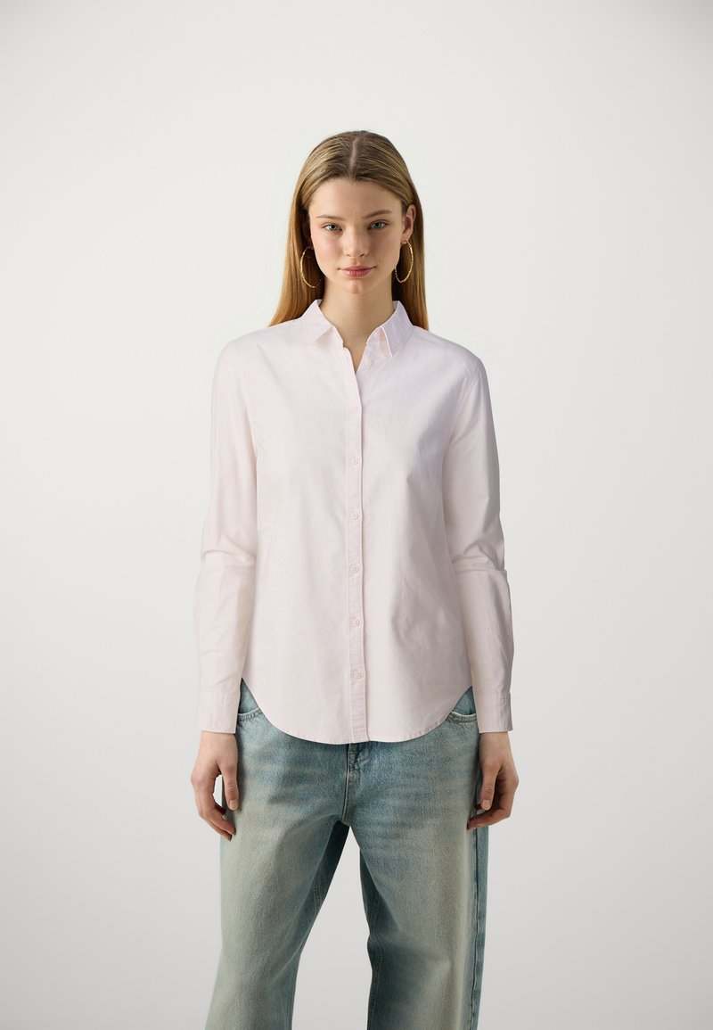 Chemise boutonnière rose clair avec un col rond et des manches longues, associée à un jean bleu clair, ample et décontracté. Design minimaliste.