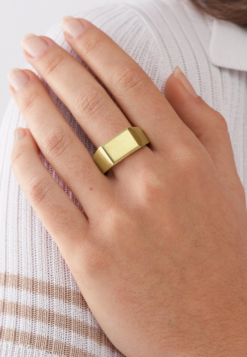 Anello in ottone con un design geometrico, caratterizzato da una superficie piatta e bordi angolati. La mano è appoggiata su un tessuto di colore chiaro e texture.